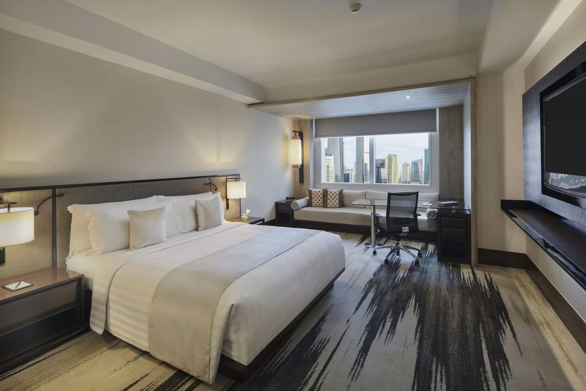 A hotel room with a large bed, a desk, a chair, a TV, and a view of the cityscape.