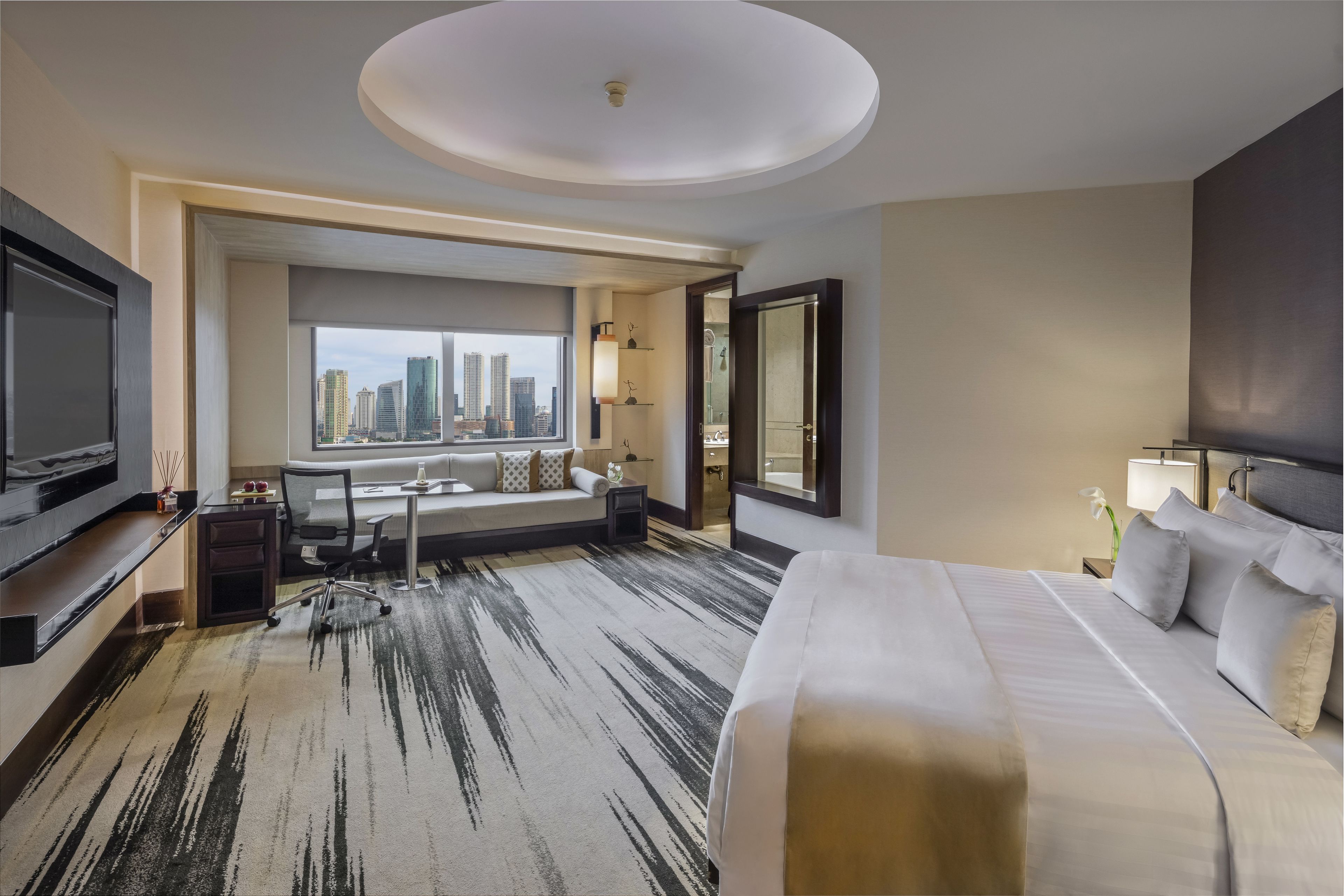 A hotel room with a large bed, a TV, a sofa, a desk, and a view of the city skyline.