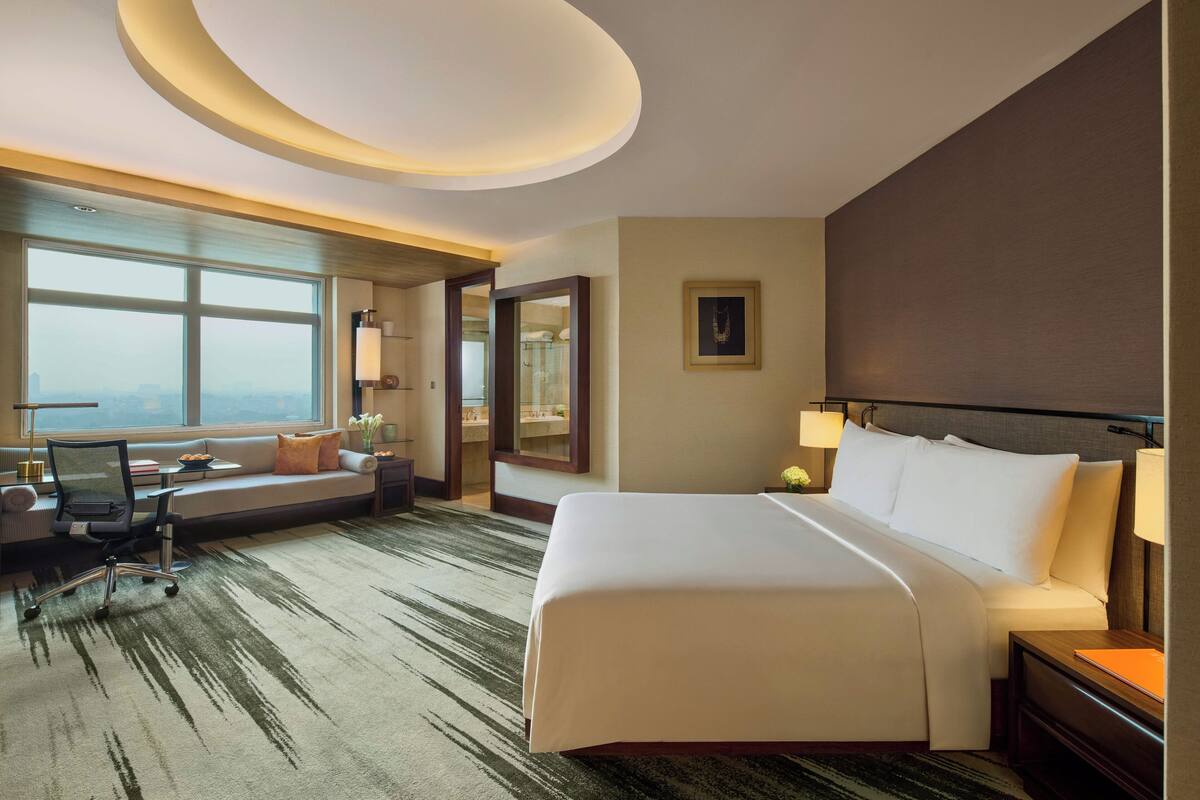 A hotel room with a large bed, a desk with a chair, a sofa, a large window, and a bathroom visible through an open door.