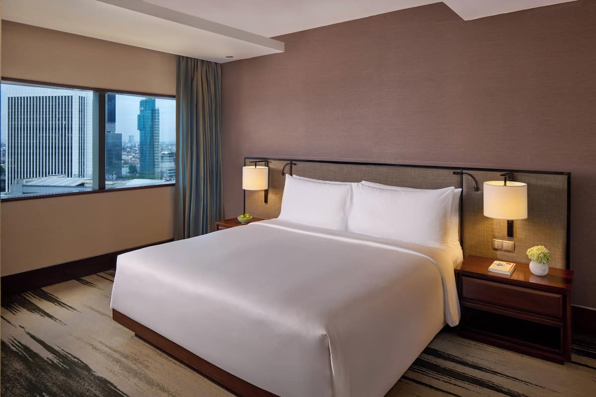 A hotel room with a large bed, two bedside lamps, a nightstand, and a view of a cityscape through large windows.