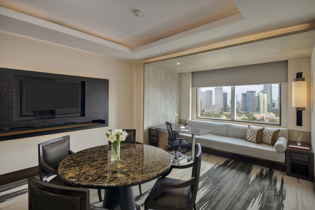 A modern hotel room with a round dining table, a sofa with a city view, a flat-screen TV mounted on the wall, and a large window overlooking the cityscape.