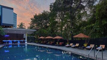A swimming pool with lounge chairs and umbrellas, surrounded by trees and a modern building.