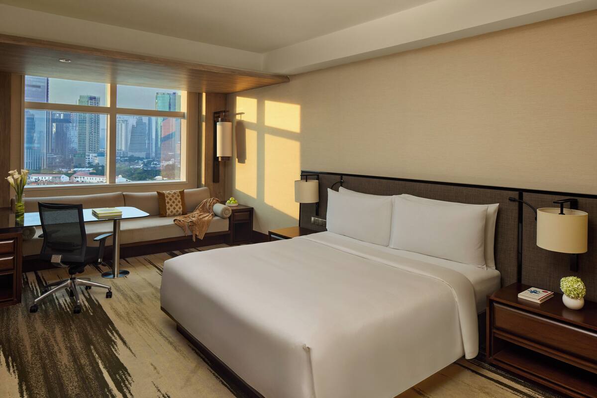 A hotel room with a large bed, a desk, a chair, a lamp, and a view of the cityscape.