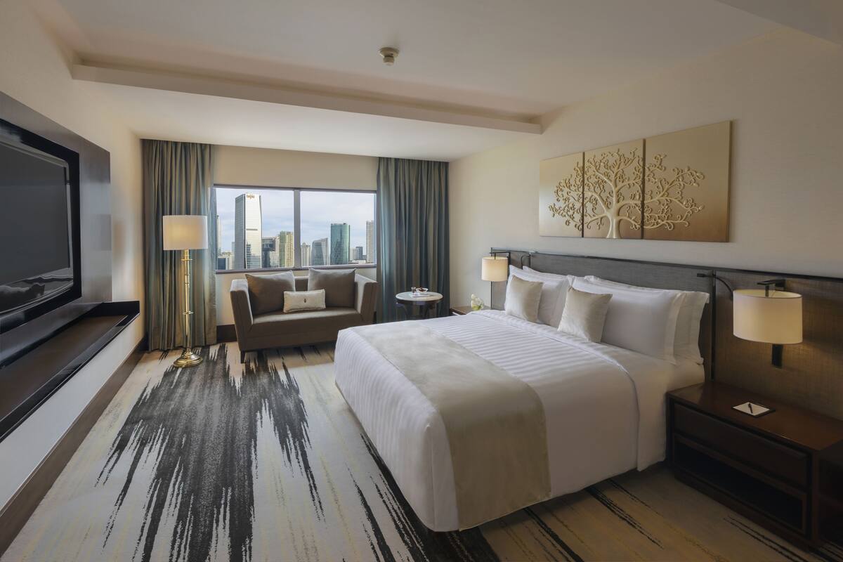 A hotel room with a large bed, a sofa, a TV, a city view, and a decorative tree artwork on the wall.