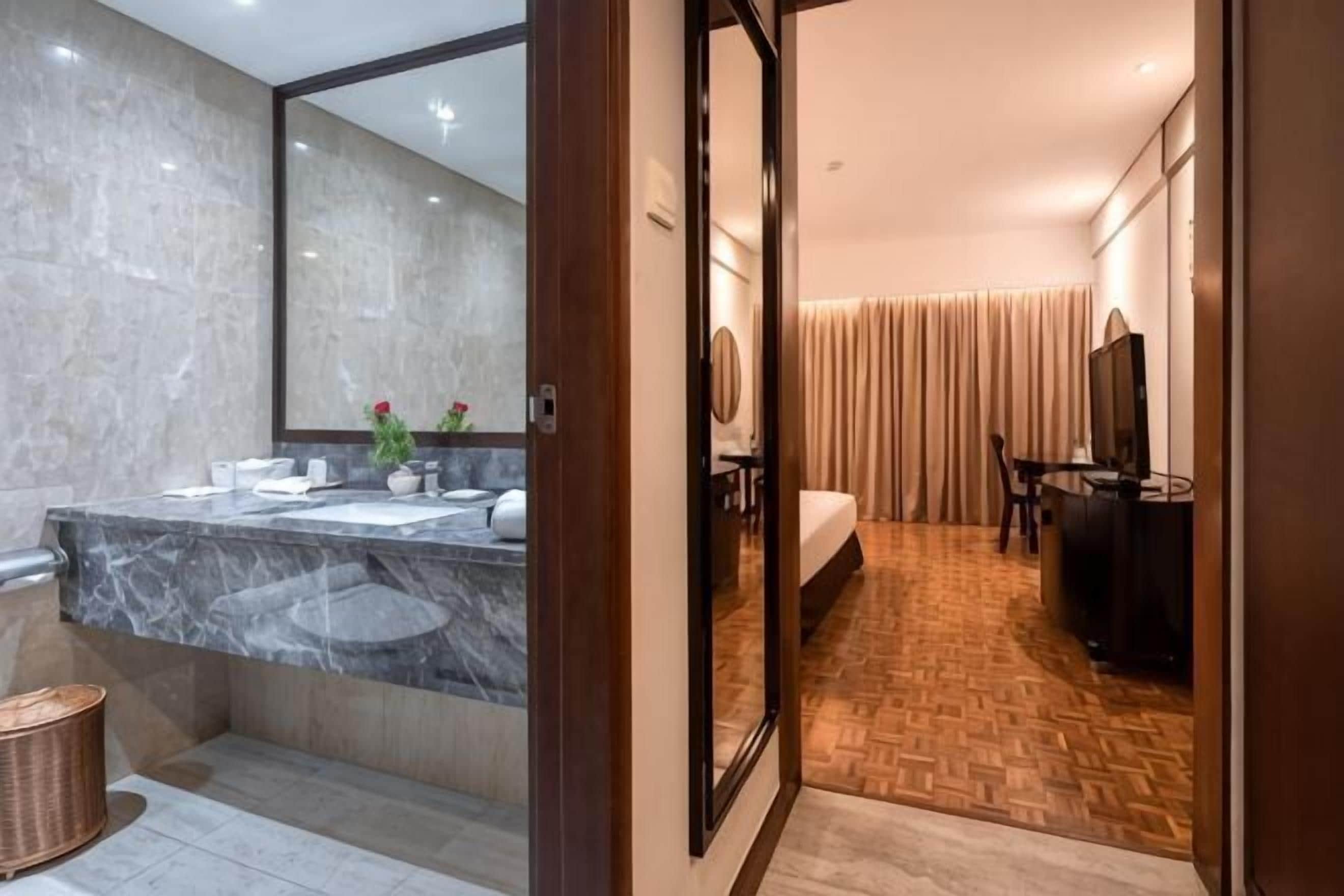 A modern bathroom with a marble sink, a large mirror, and a wooden floor leading to a bedroom with a TV and curtains.