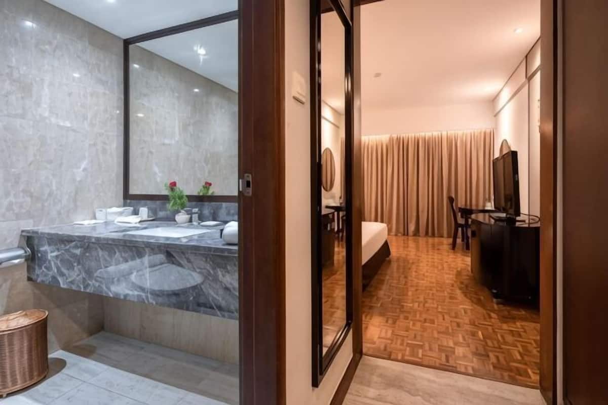 A modern bathroom with a marble sink, a large mirror, and a wooden floor leading to a bedroom with a TV and curtains.