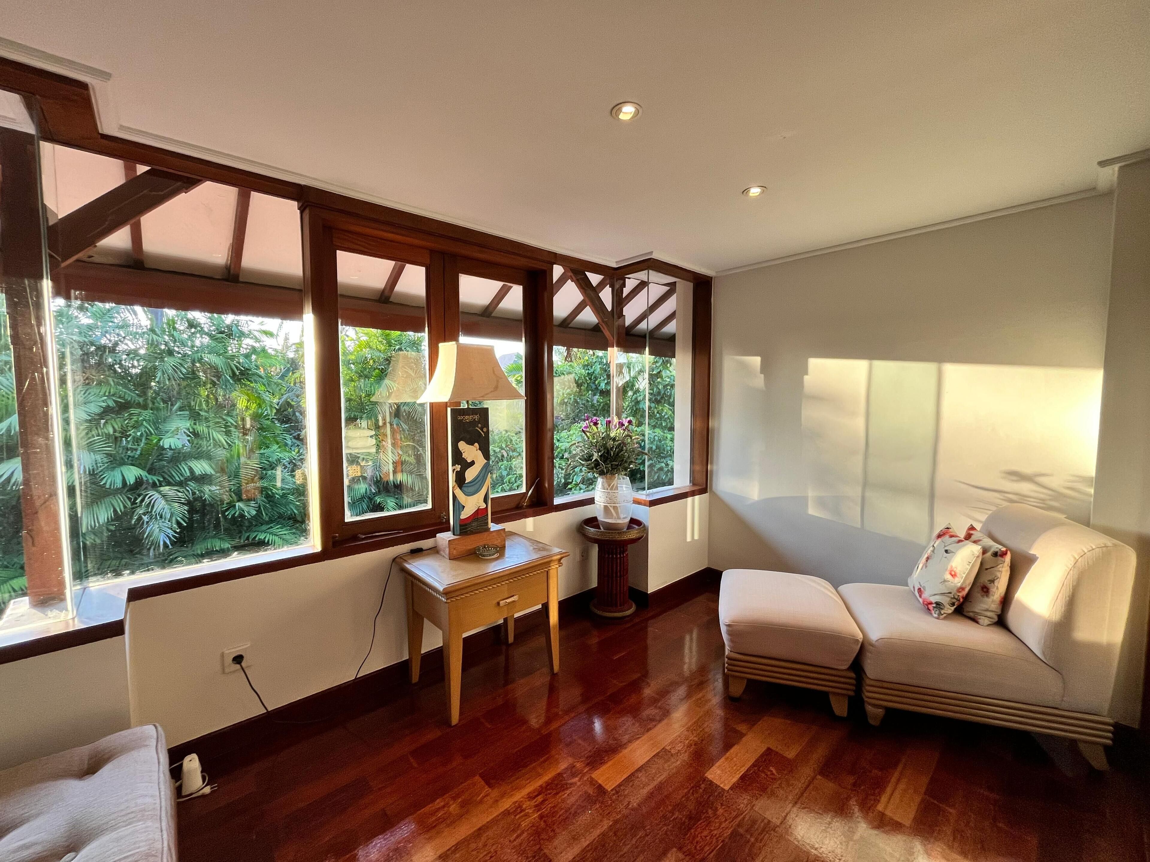 A room with a wooden floor, a sofa, a table with a lamp, and a window with a view of greenery.