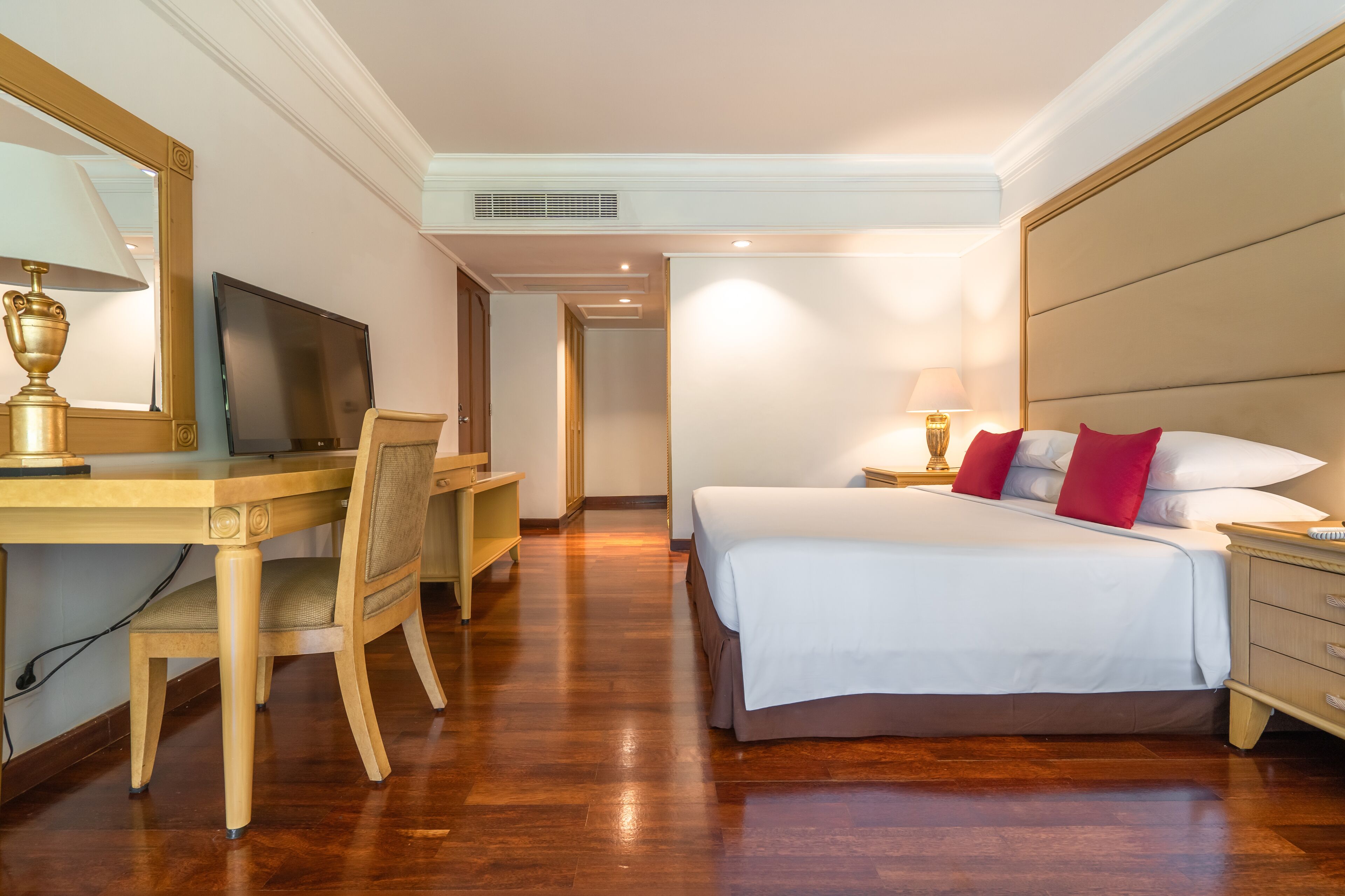 Habitación de hotel con un escritorio de madera, televisión, cama con almohadas blancas y rojas, y una mesita de noche.