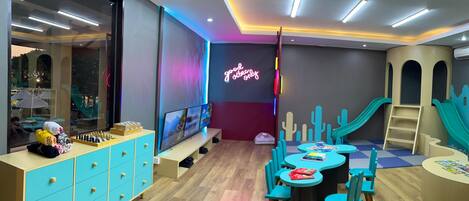 Una sala de juegos para niños con un letrero de neón, zona de recreación y una televisión grande.