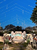 An outdoor wedding venue with a decorated stage, string lights, and floral arrangements.
