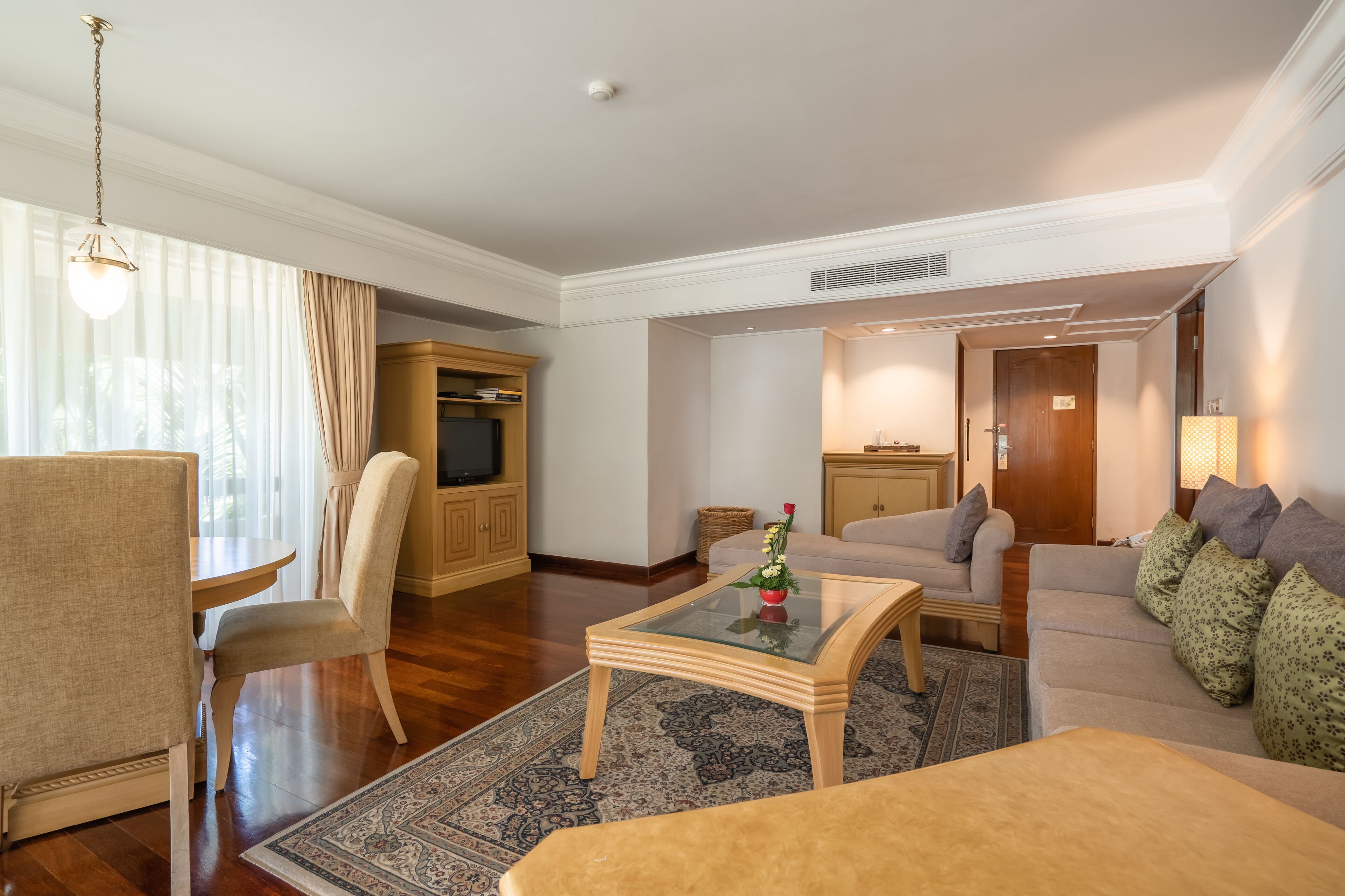 A spacious living room with a sofa, armchair, and a glass-top coffee table. There is a television in a wooden cabinet, and a lamp on a side table.
