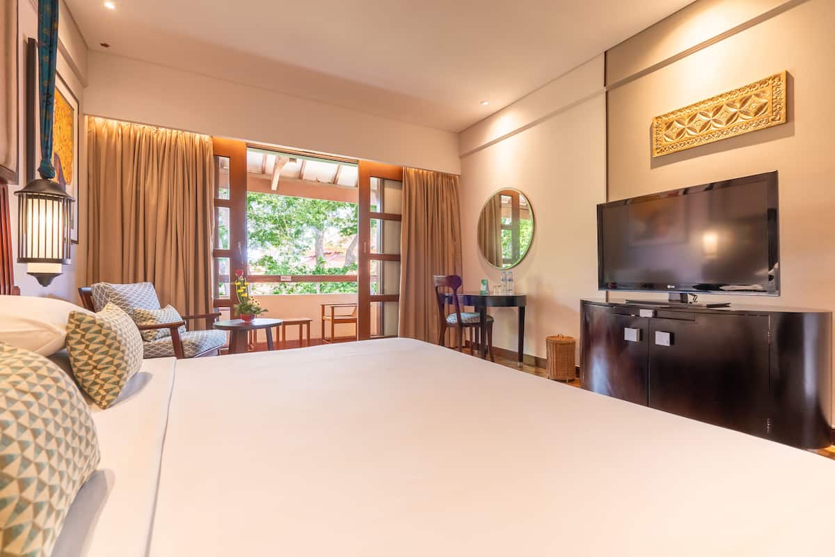 A hotel room with a large bed, a TV, a desk, and a view of greenery outside.