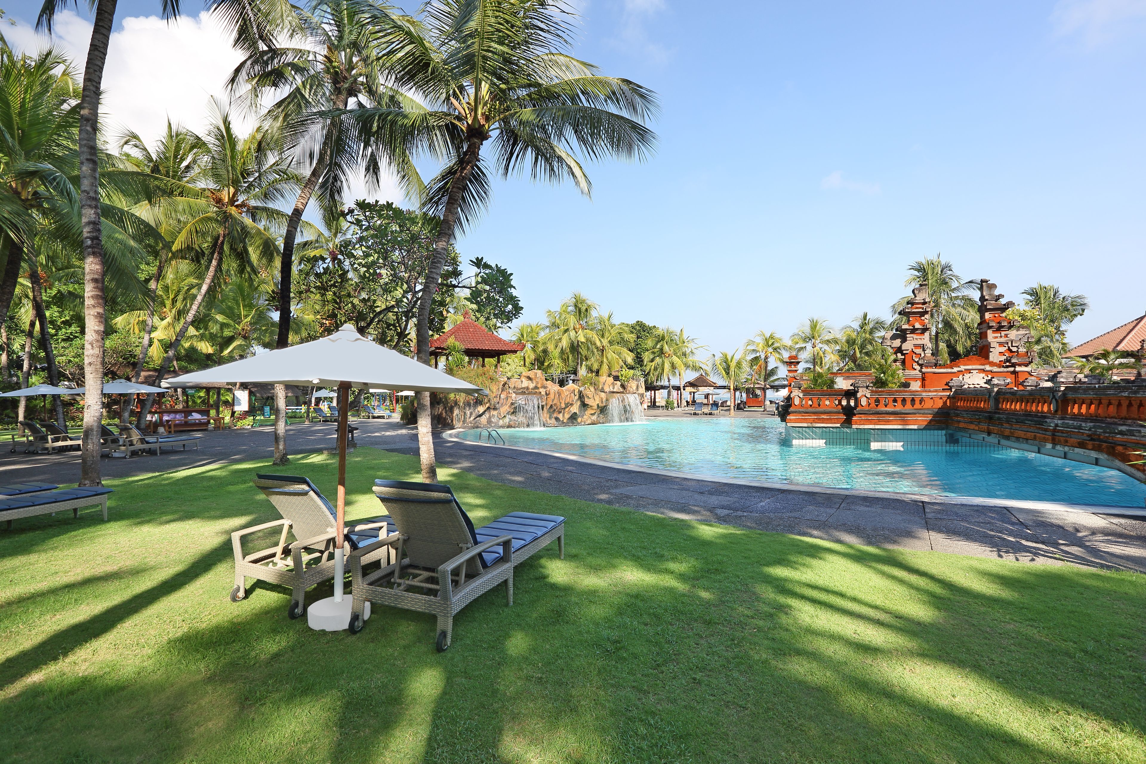 A pool area with lounge chairs and umbrellas, surrounded by palm trees and a traditional building.