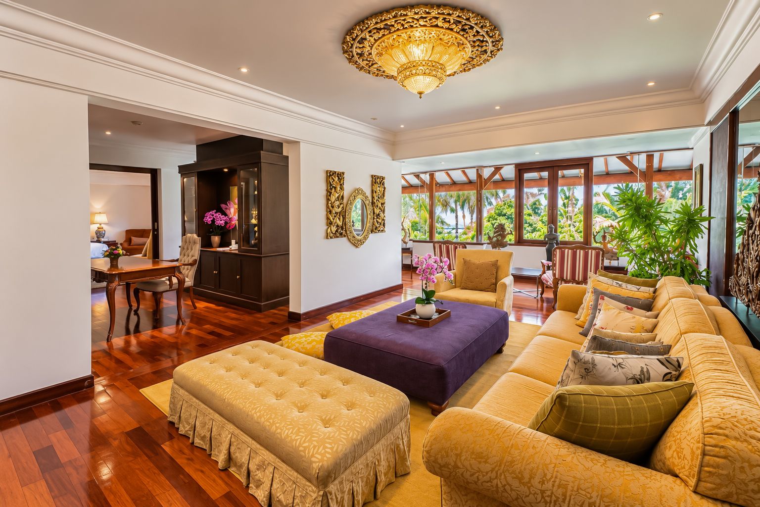 A spacious living area with a large sofa, a coffee table, and a chandelier. There is a dining area with a table and chairs, and a view of greenery through large windows.