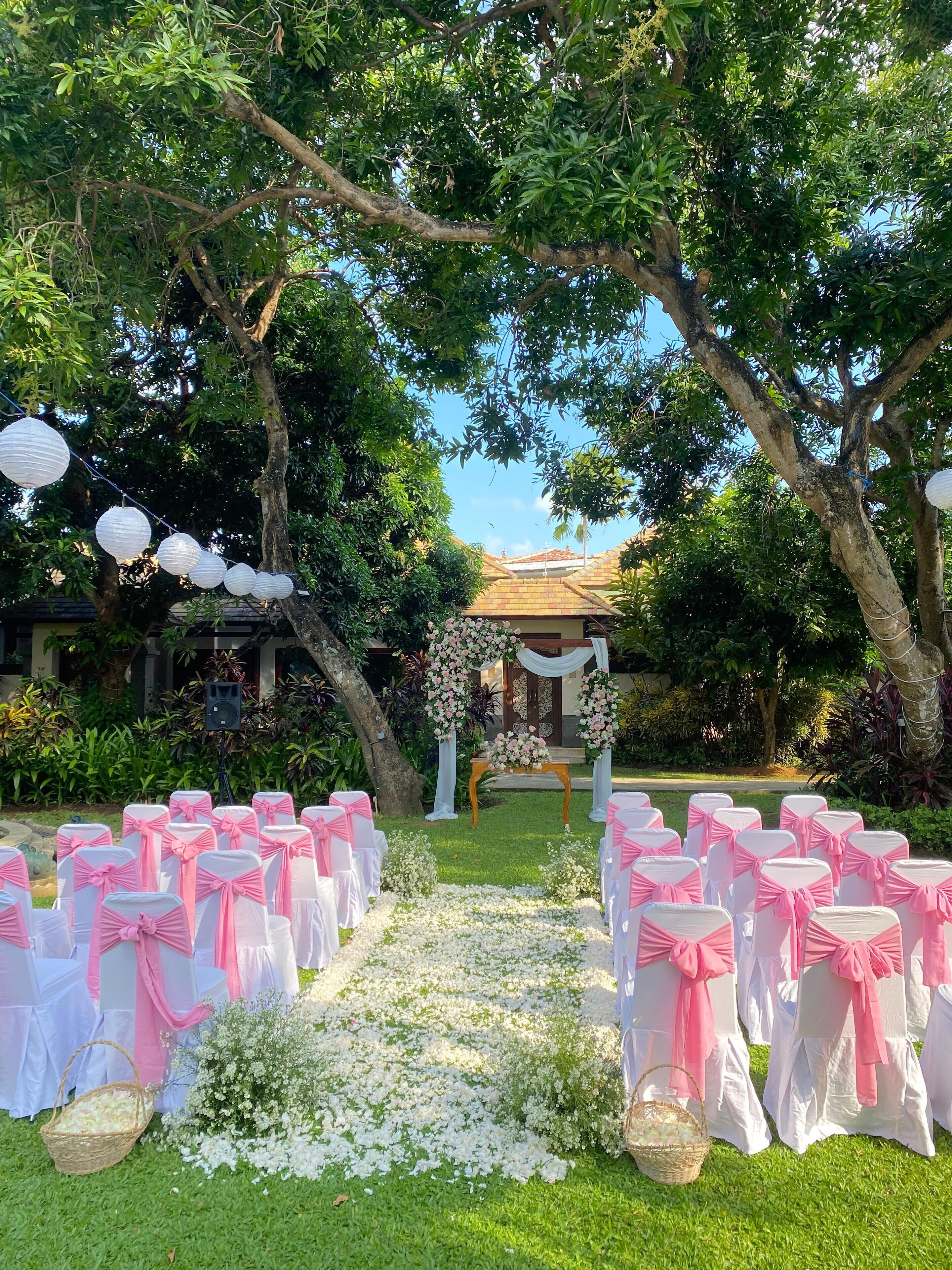 A wedding ceremony setup with a floral arch, white and pink chairs, and a speaker.