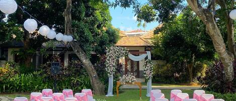A wedding ceremony setup with a floral arch, white and pink chairs, and a speaker.