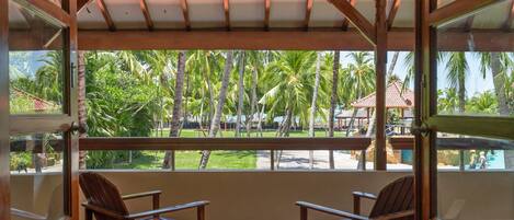 Un área de estar al aire libre cubierta con sillas de madera y una mesa, con vistas a un paisaje tropical con palmeras y una piscina.