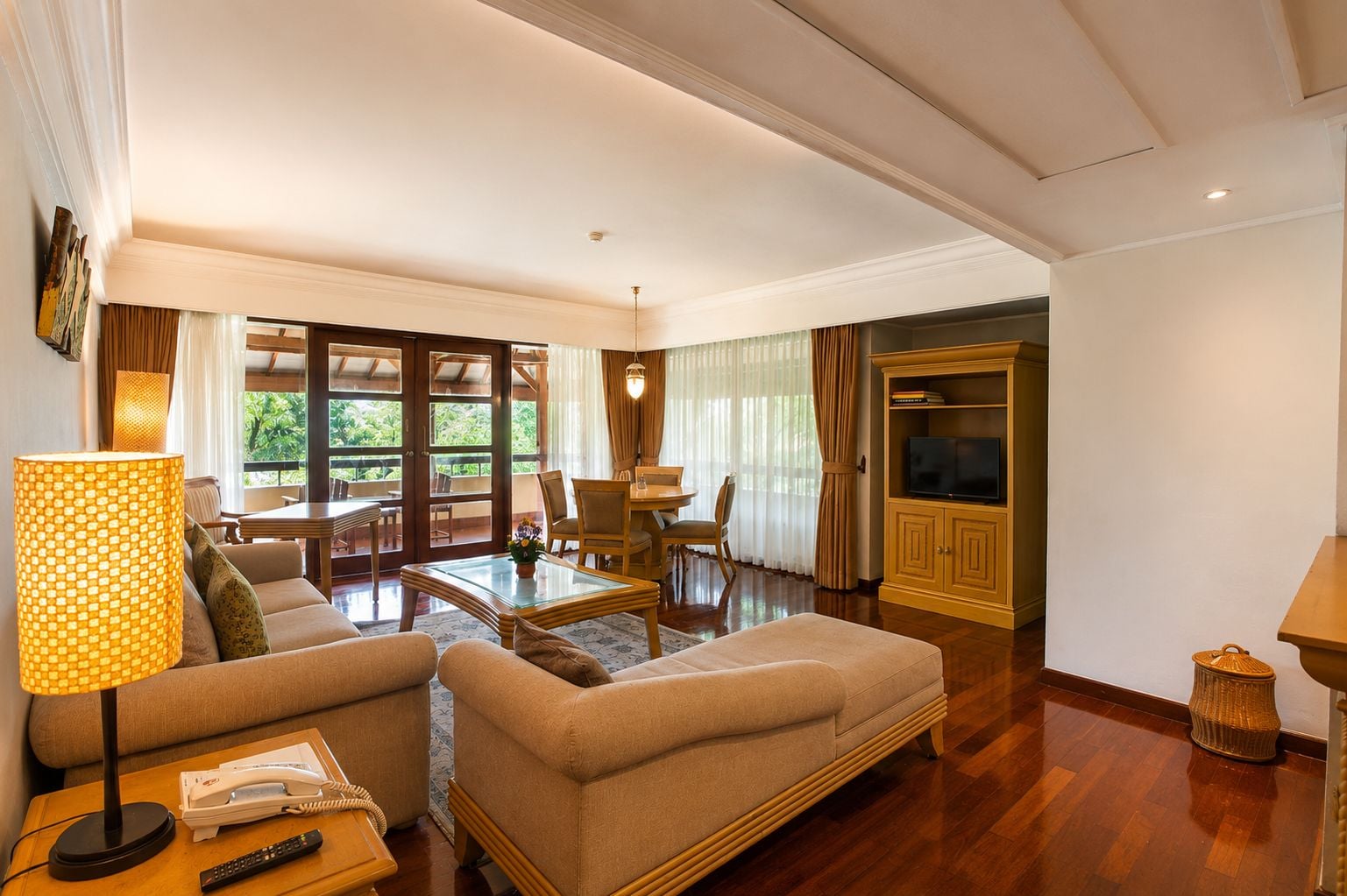 A spacious living room with a sofa, armchair, and a dining area with a table and chairs. A television is mounted on a cabinet, and there is a lamp on a side table.