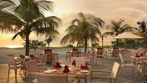 An outdoor dining area with wicker furniture, tables set with red napkins, and palm trees.