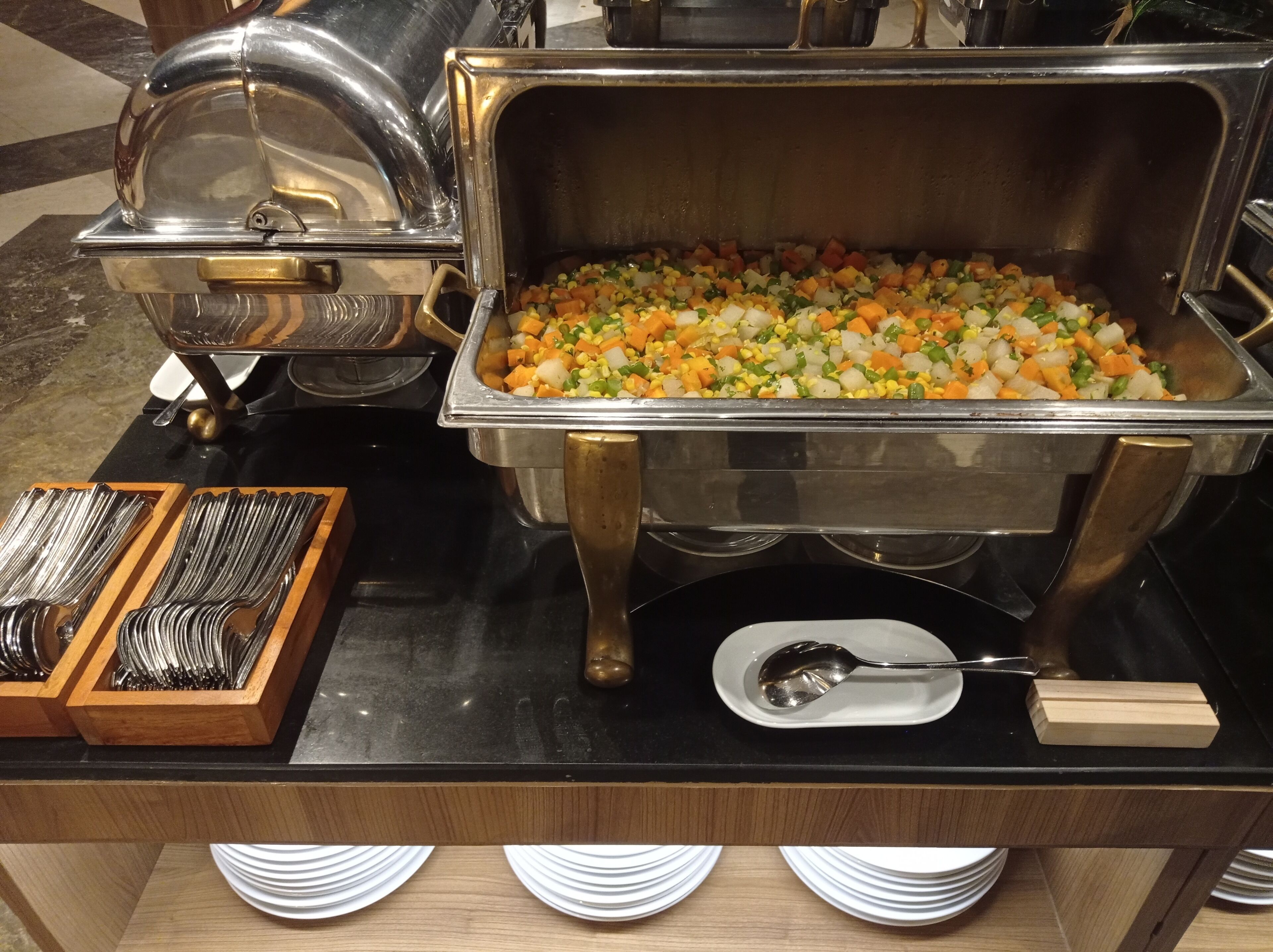 A buffet setup with a variety of vegetables in a large metal container, accompanied by serving utensils and plates.