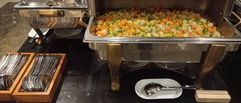 A buffet setup with a variety of vegetables in a large metal container, accompanied by serving utensils and plates.