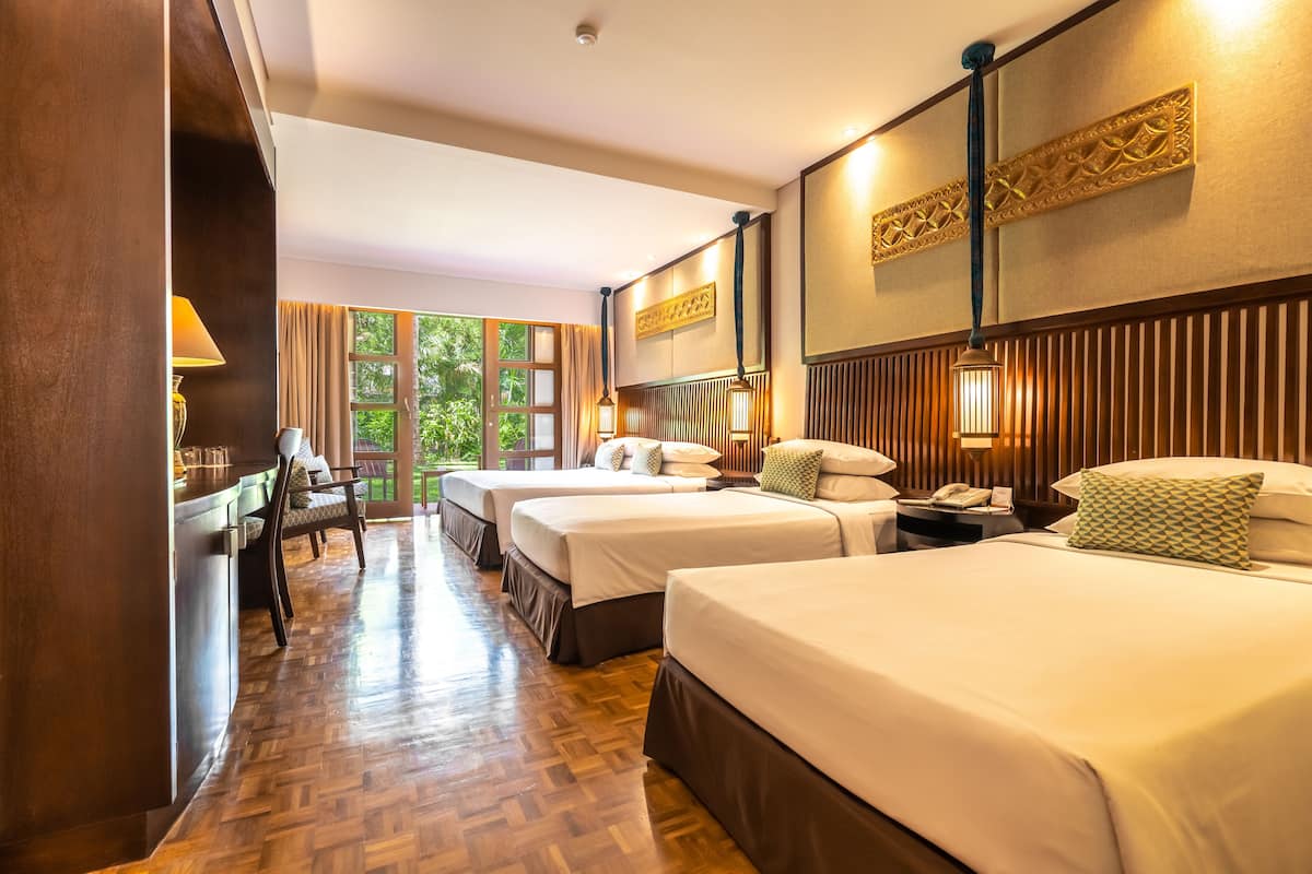A hotel room with two beds, a desk, and a chair. There is a window with a view of greenery outside.