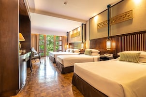 A hotel room with two beds, a desk, and a chair. There is a window with a view of greenery outside.