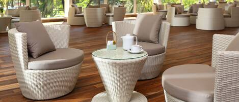 A spacious indoor area with wicker furniture and a glass coffee table, featuring a view of lush greenery outside.