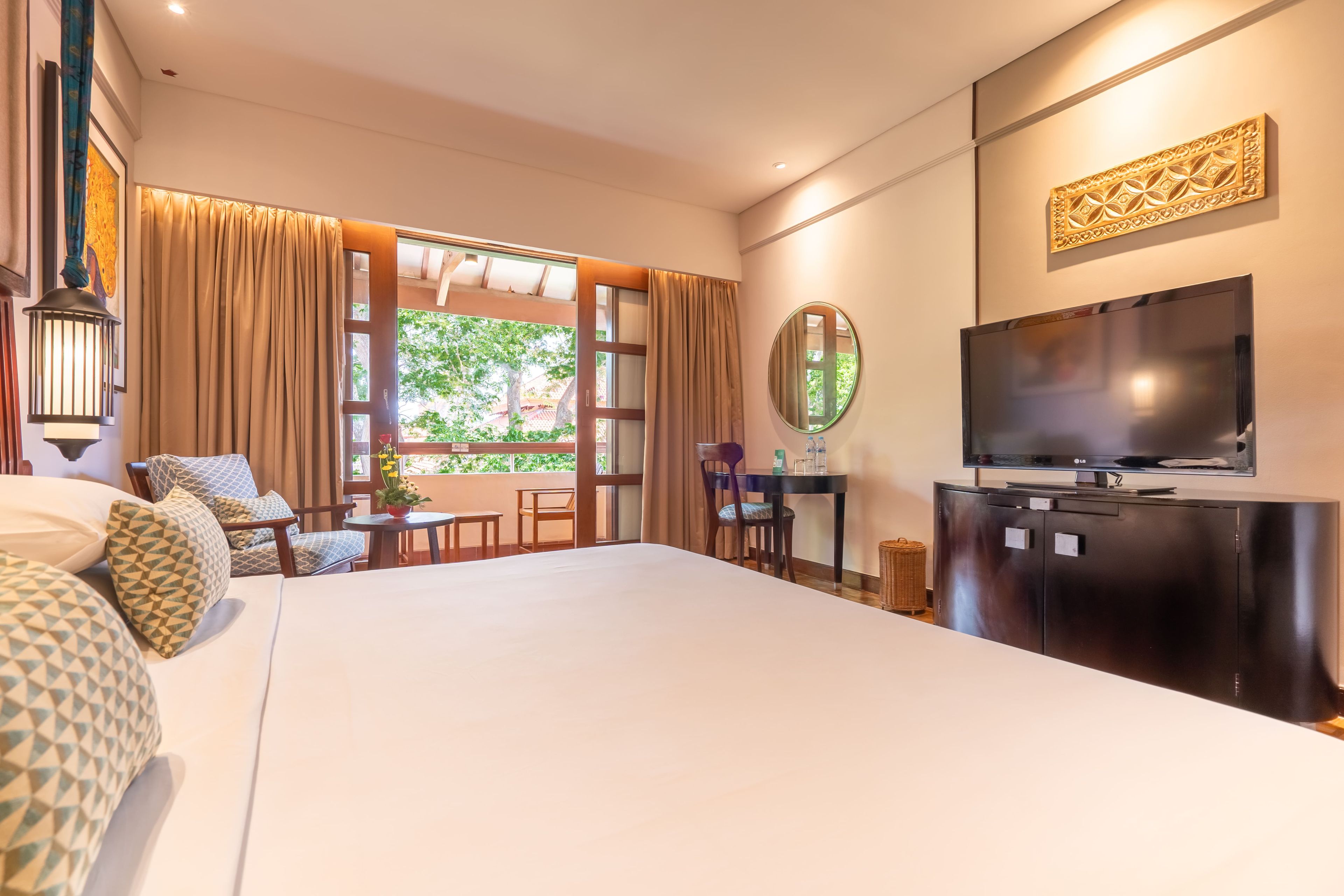 A hotel room with a large bed, a TV, a dining table, and a view of greenery outside.