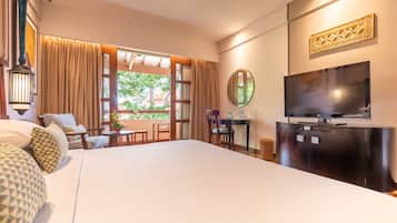 A hotel room with a large bed, a TV, a dining table, and a view of greenery outside.