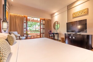 A hotel room with a large bed, a TV, a dining table, and a view of greenery outside.