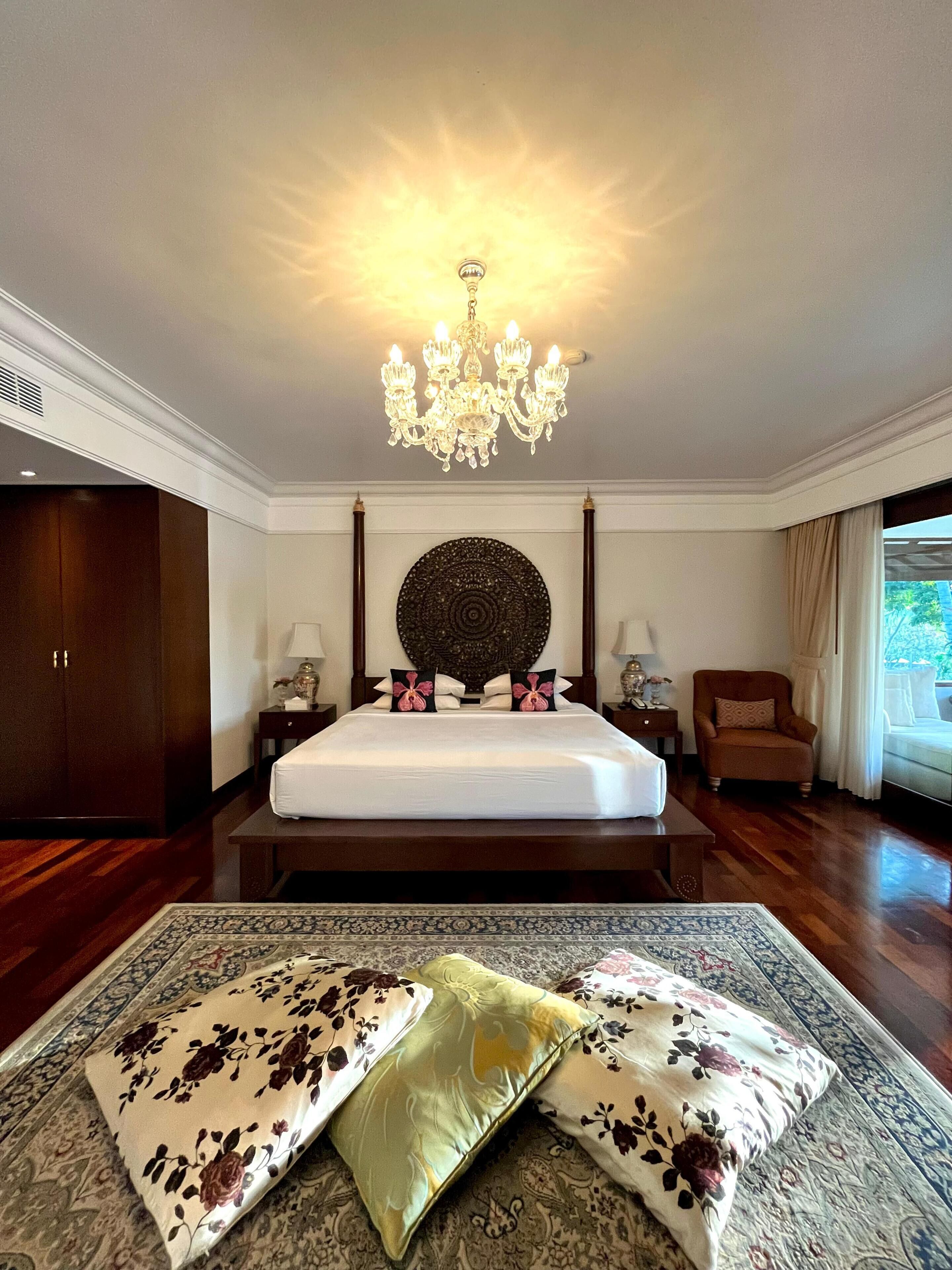 A hotel room with a large bed, a chandelier, a wooden wardrobe, a sitting area with a chair, and a view of the outdoors.