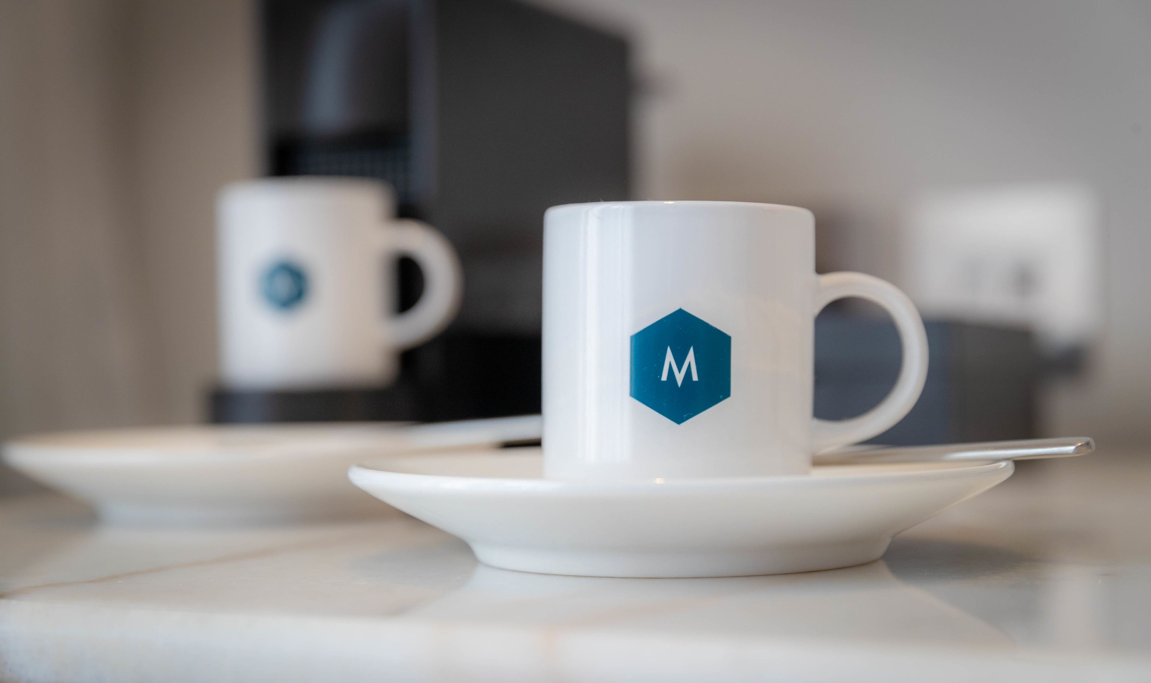 Una taza de café blanca con un logo azul sobre un plato, sobre una mesa.