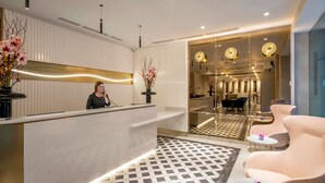 Zona de recepción de hotel con recepcionista, un mostrador de recepción, un mostrador de registro, suelo estampado y un área de estar con mesas y sillas.