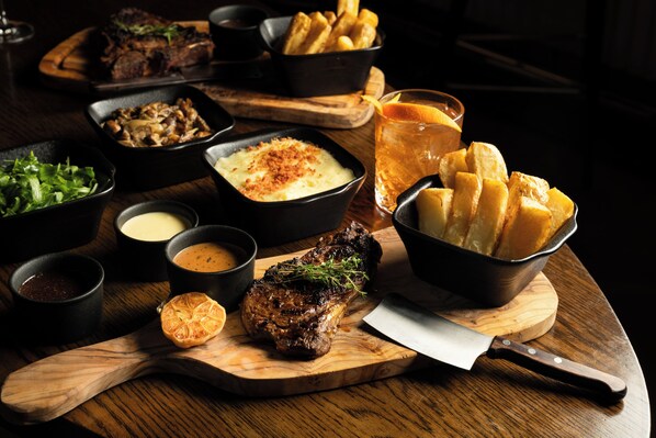 A wooden cutting board with a steak, a knife, and a bowl of fries.