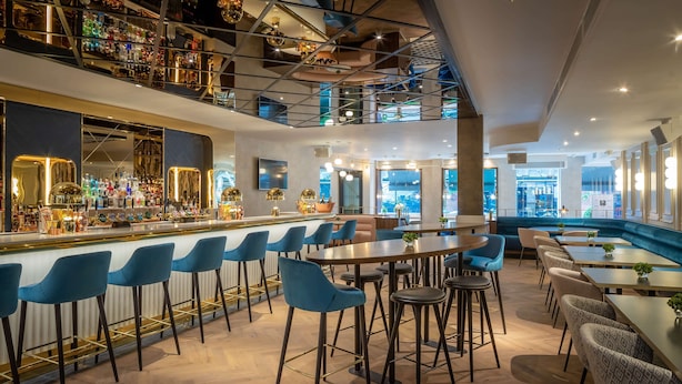 Un área de bar moderna con techo alto, taburetes azules y una variedad de botellas y copas en las estanterías.