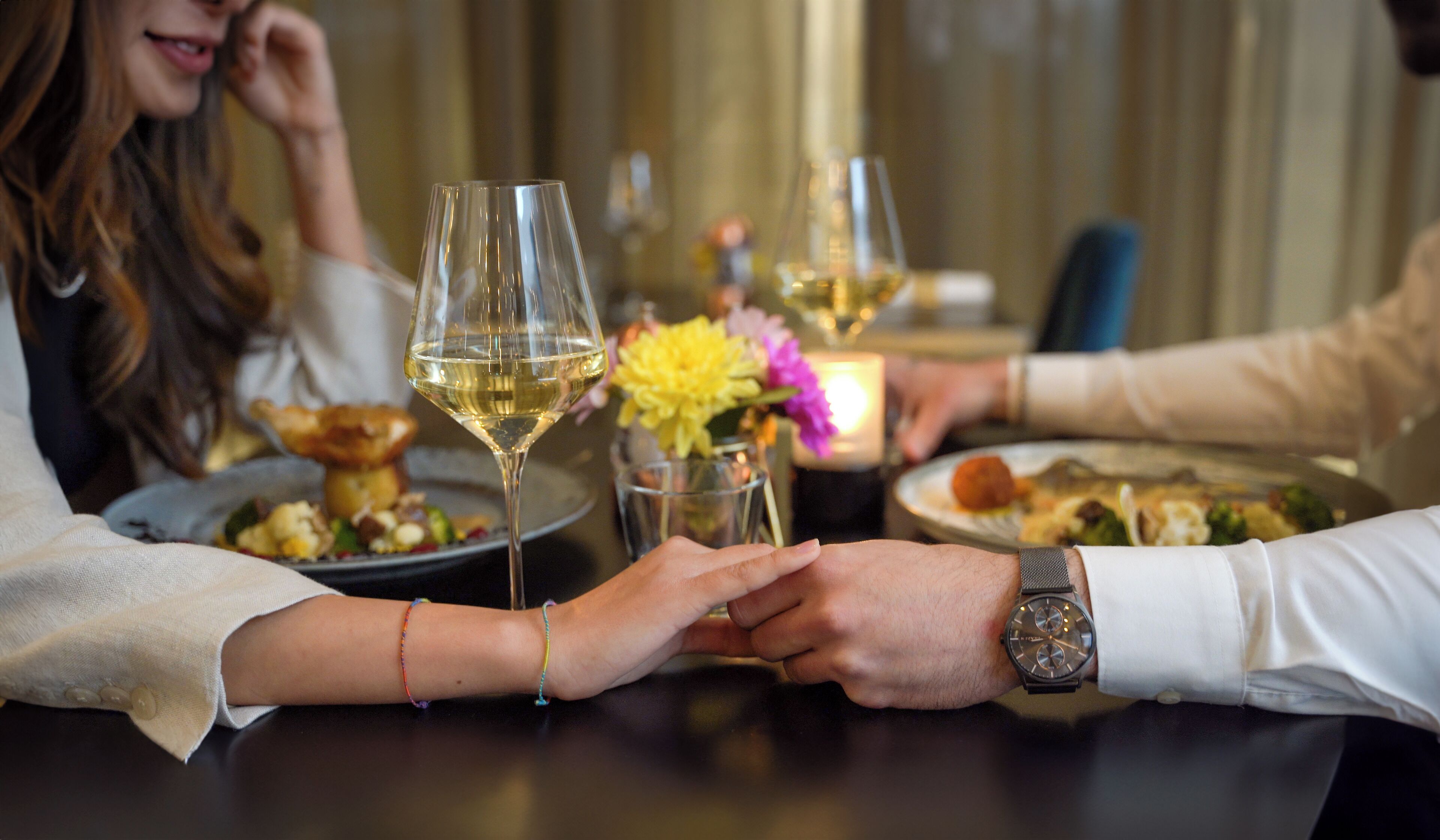 A dining table with a glass of white wine, a candle, and a vase with flowers.