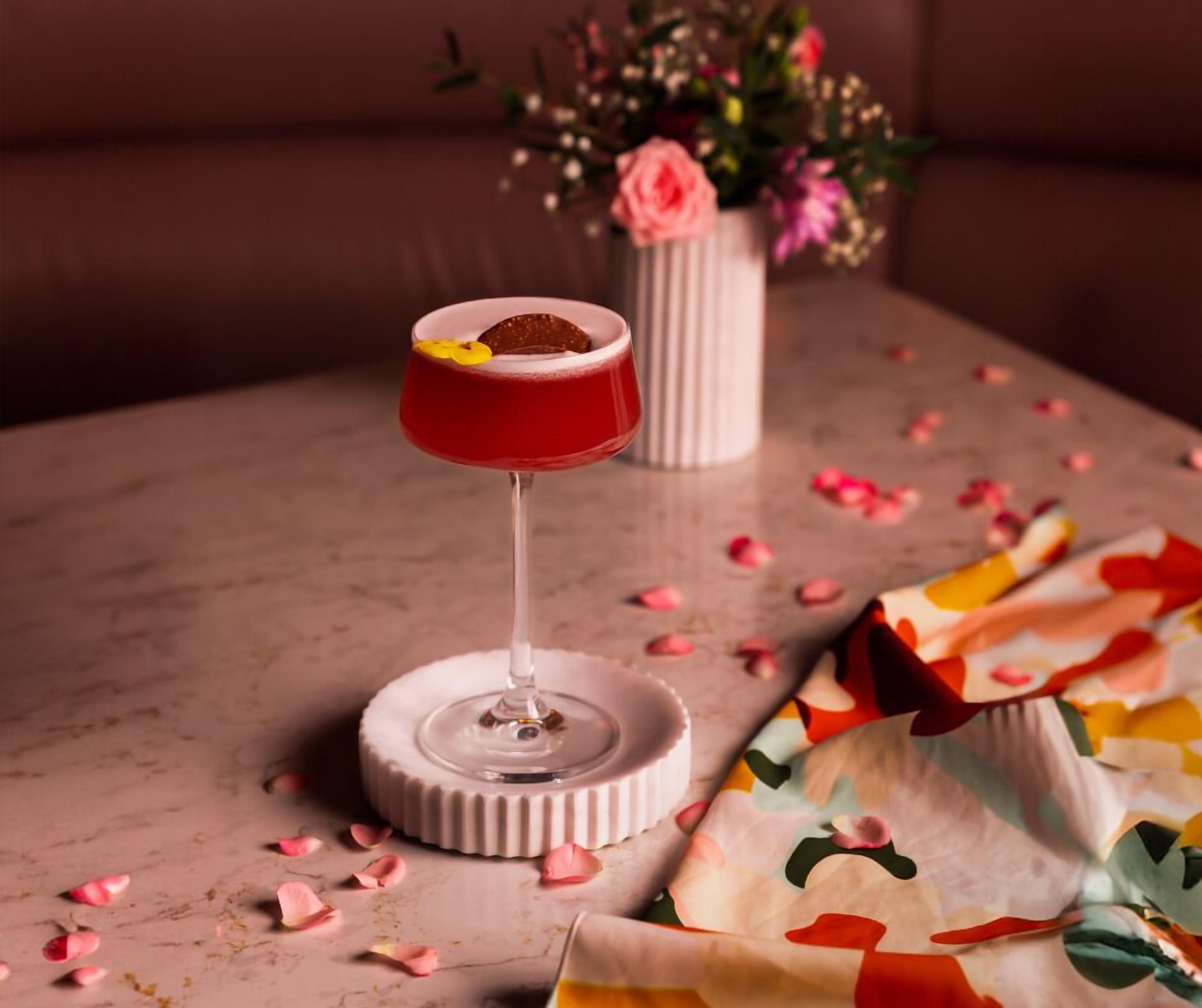 A cocktail in a red glass on a marble coaster, with rose petals scattered around.
