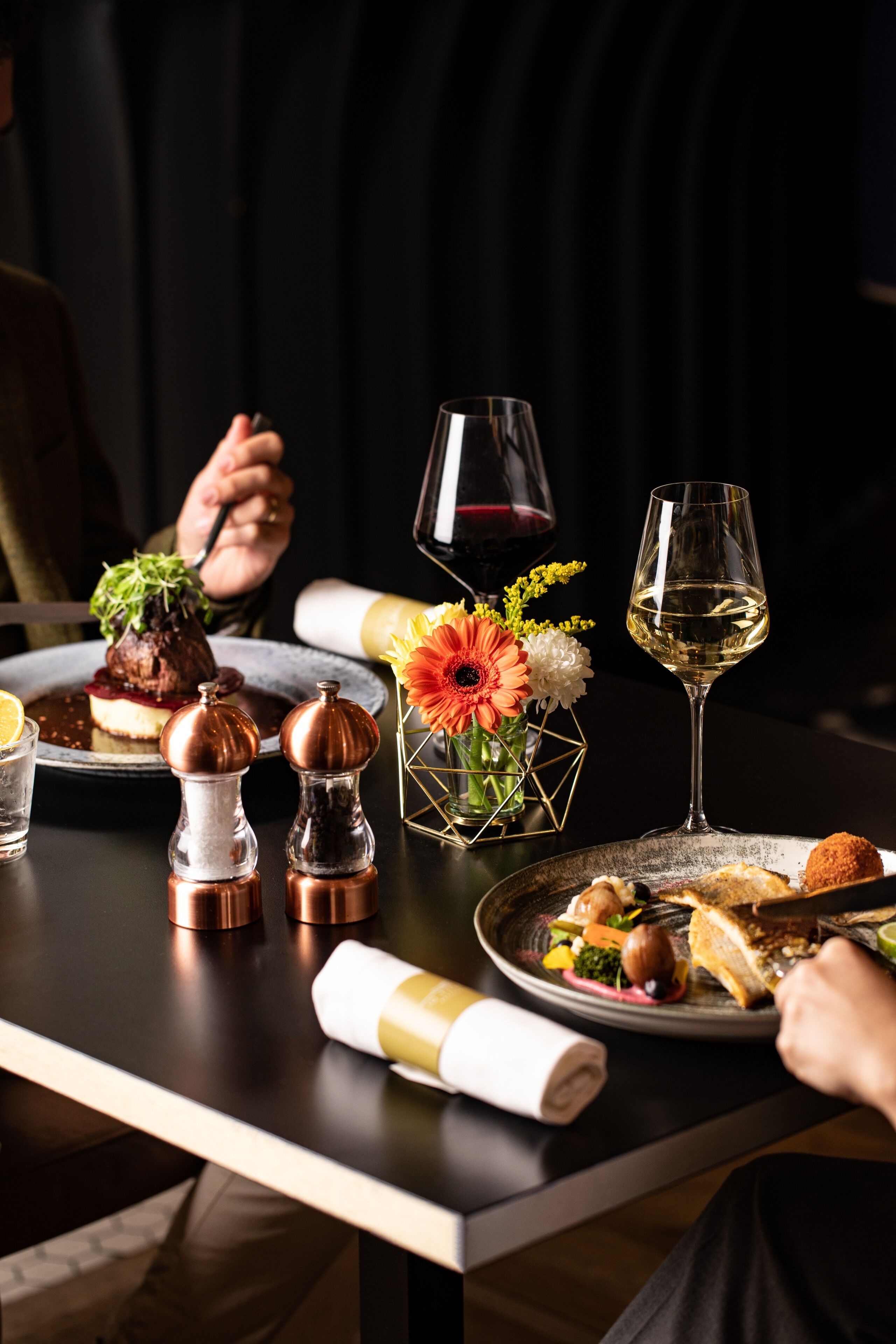 Una mesa de comedor dispuesta con platos de comida, copas de vino y flores decorativas.