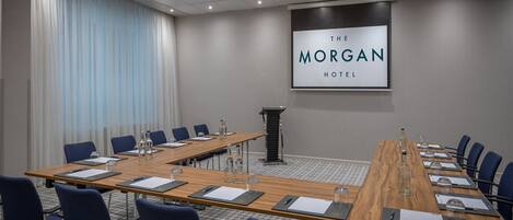 Un salón de conferencias con una larga mesa de madera, sillas azules y una pantalla proyectora mostrando "The Morgan Hotel".