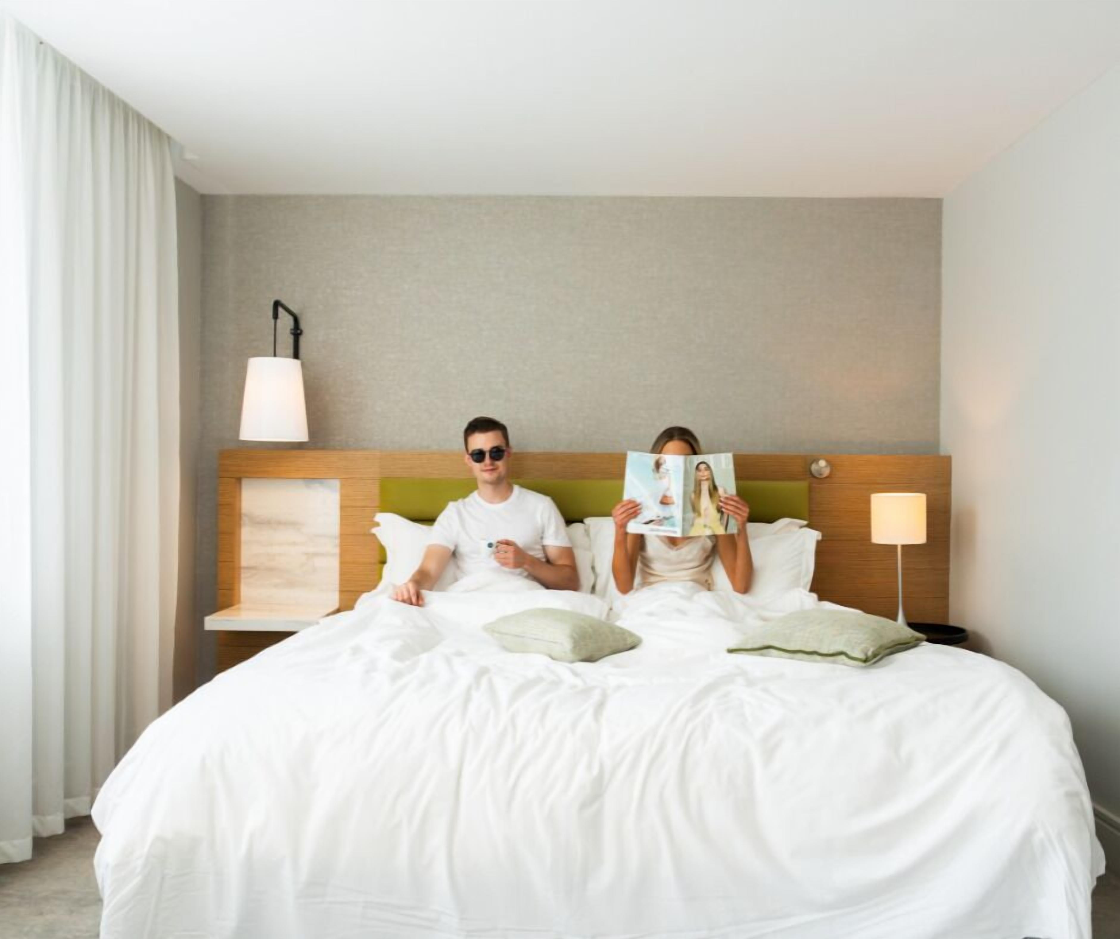 Habitación de hotel con una cama grande, una mesita de noche con lámpara y vistas al exterior.