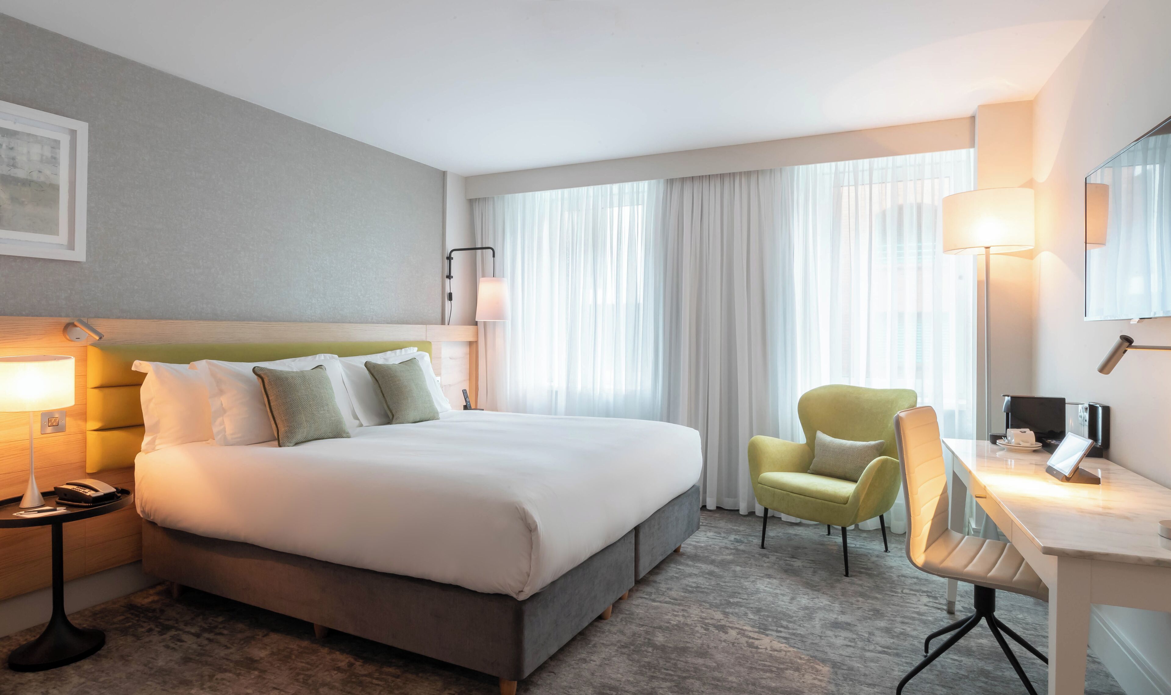 A hotel room with a bed, two chairs, a desk, a lamp, and a large window with curtains.
