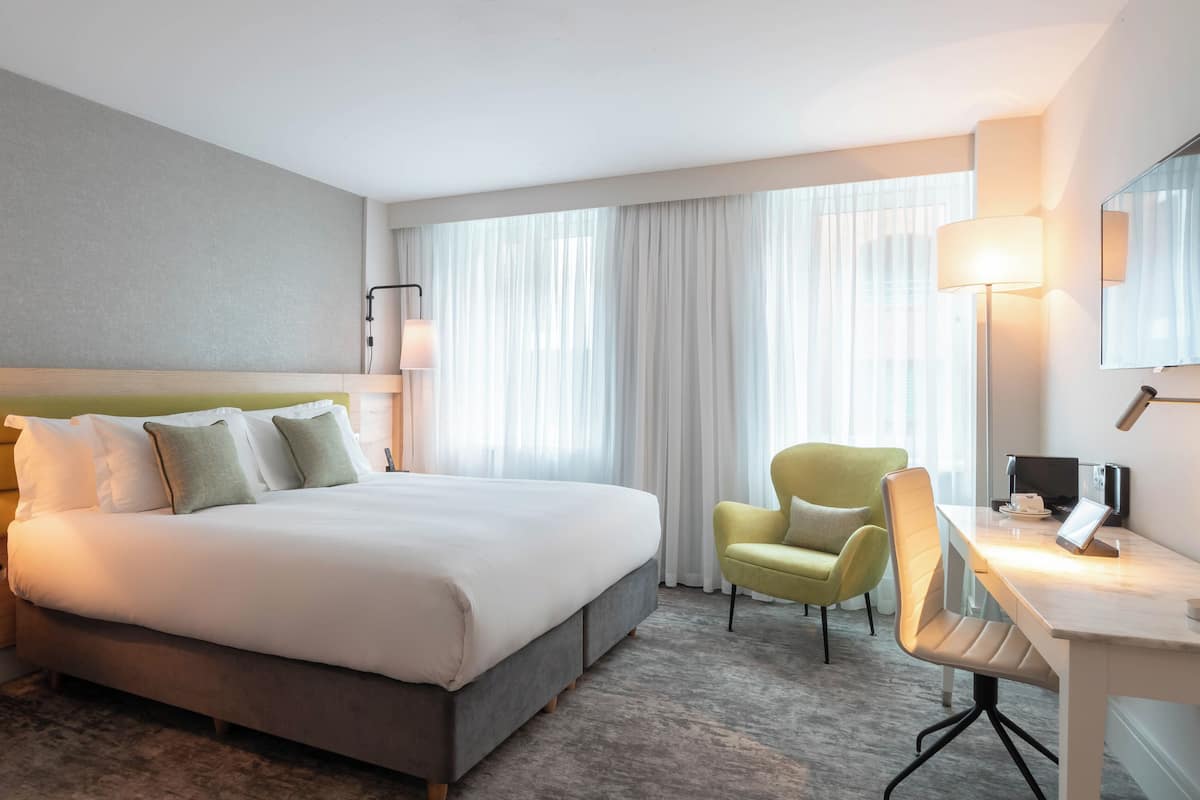 A hotel room with a bed, two chairs, a desk, a lamp, and a large window with curtains.