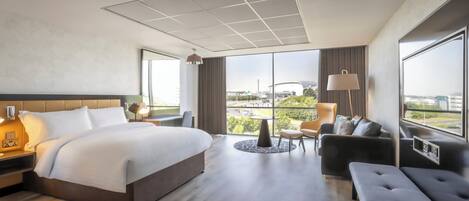 Habitación de hotel moderna con una cama grande, una zona de estar con un sofá y sillones, una mesita y vistas a la ciudad a través de amplios ventanales.