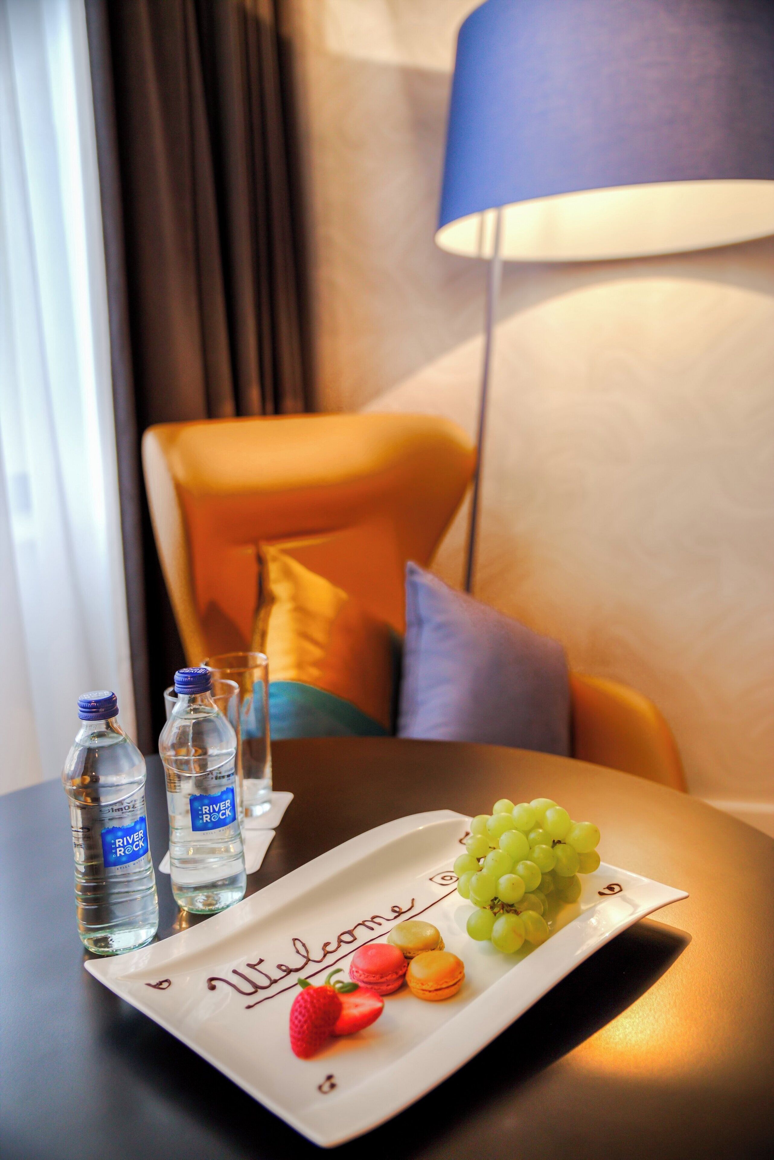 Hotelkamer met een tafel, compleet met een welkomstbord, twee waterflessen, een glas, een lamp en een fauteuil met kussens.