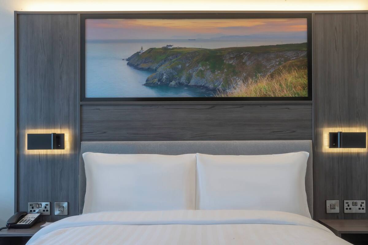 A hotel room with a large bed, a flat-screen TV mounted on the wall displaying a coastal landscape, and bedside lamps.