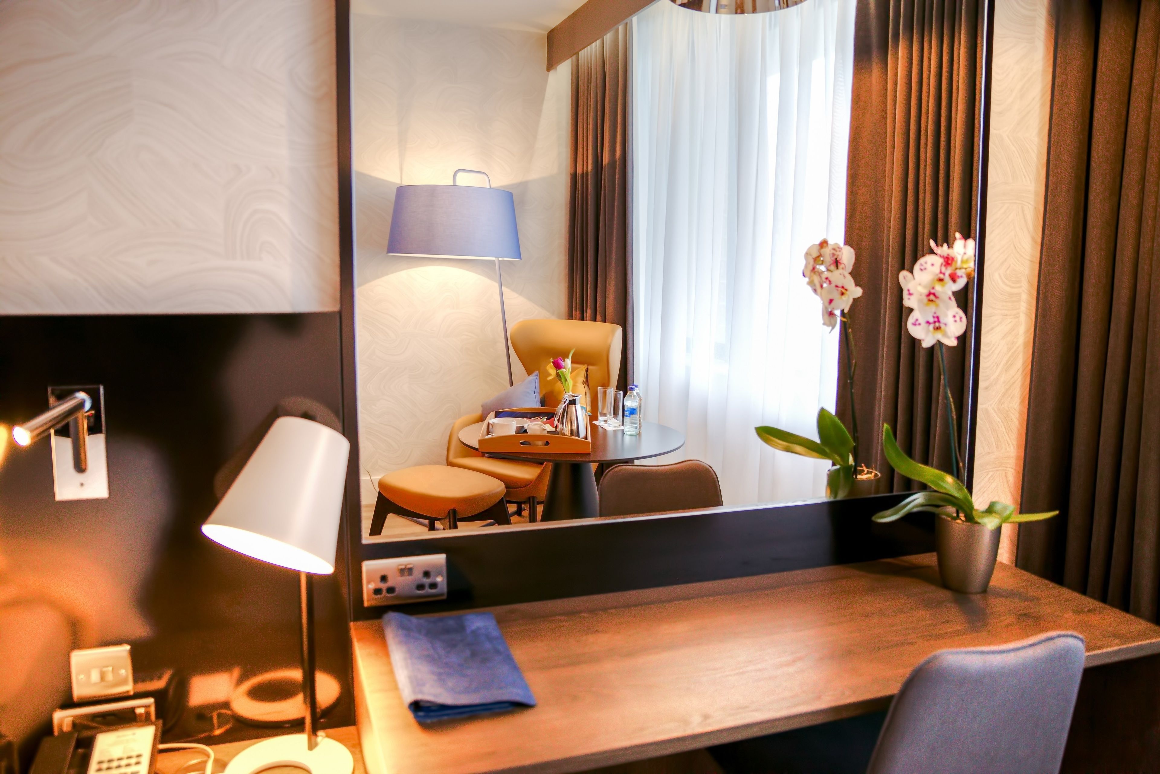 A hotel room with a wooden desk, a mirror, a lamp, a chair, a small table with a vase of flowers, and a potted plant.