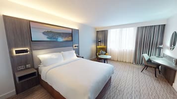 A hotel room with a large bed, a desk, a chair, a small table, a lamp, a mirror, and a painting of a coastal landscape.