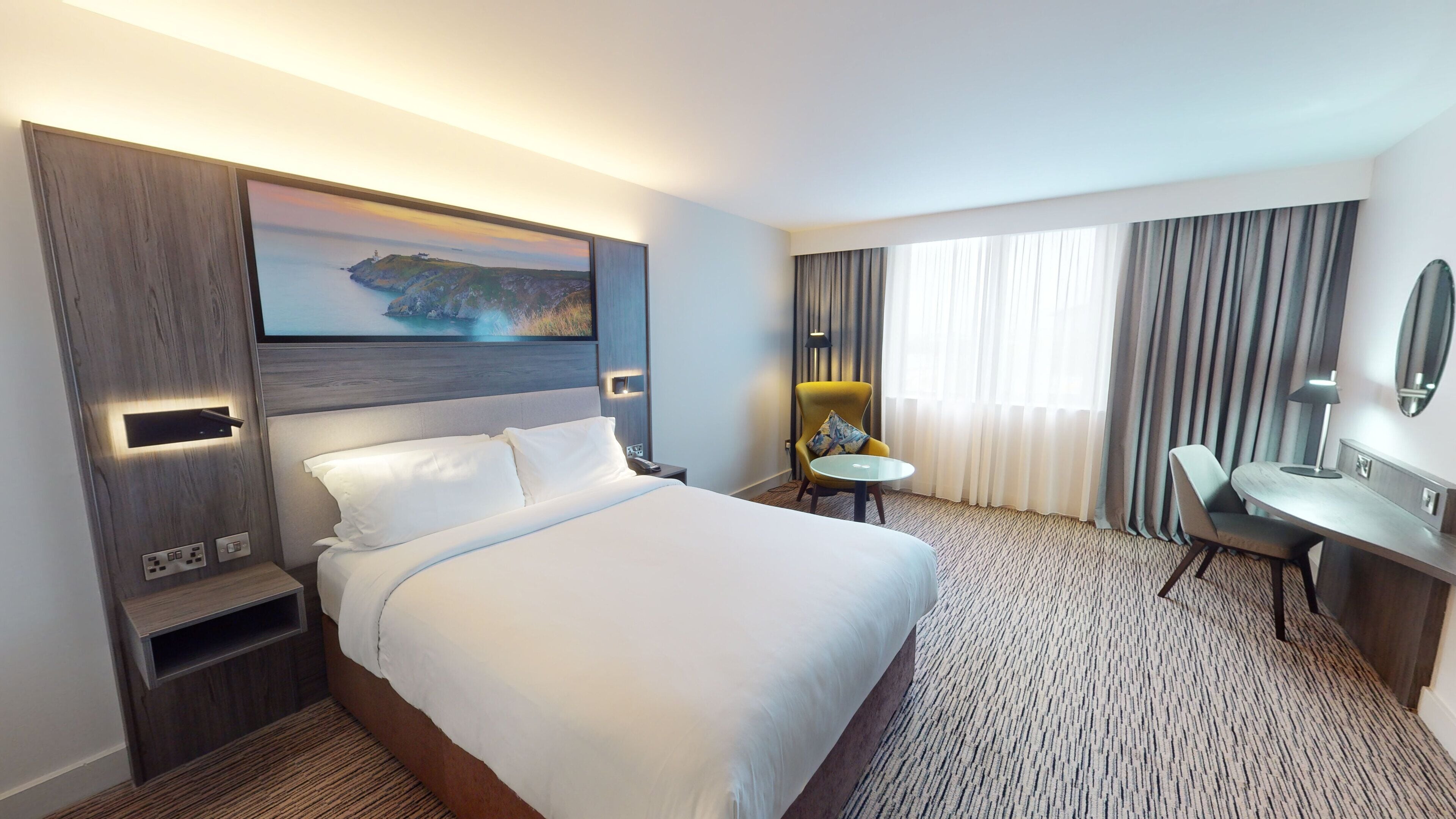 Una habitación de hotel con una cama grande, un escritorio, una silla, una mesita redonda pequeña, una lámpara y un cuadro de un paisaje costero.