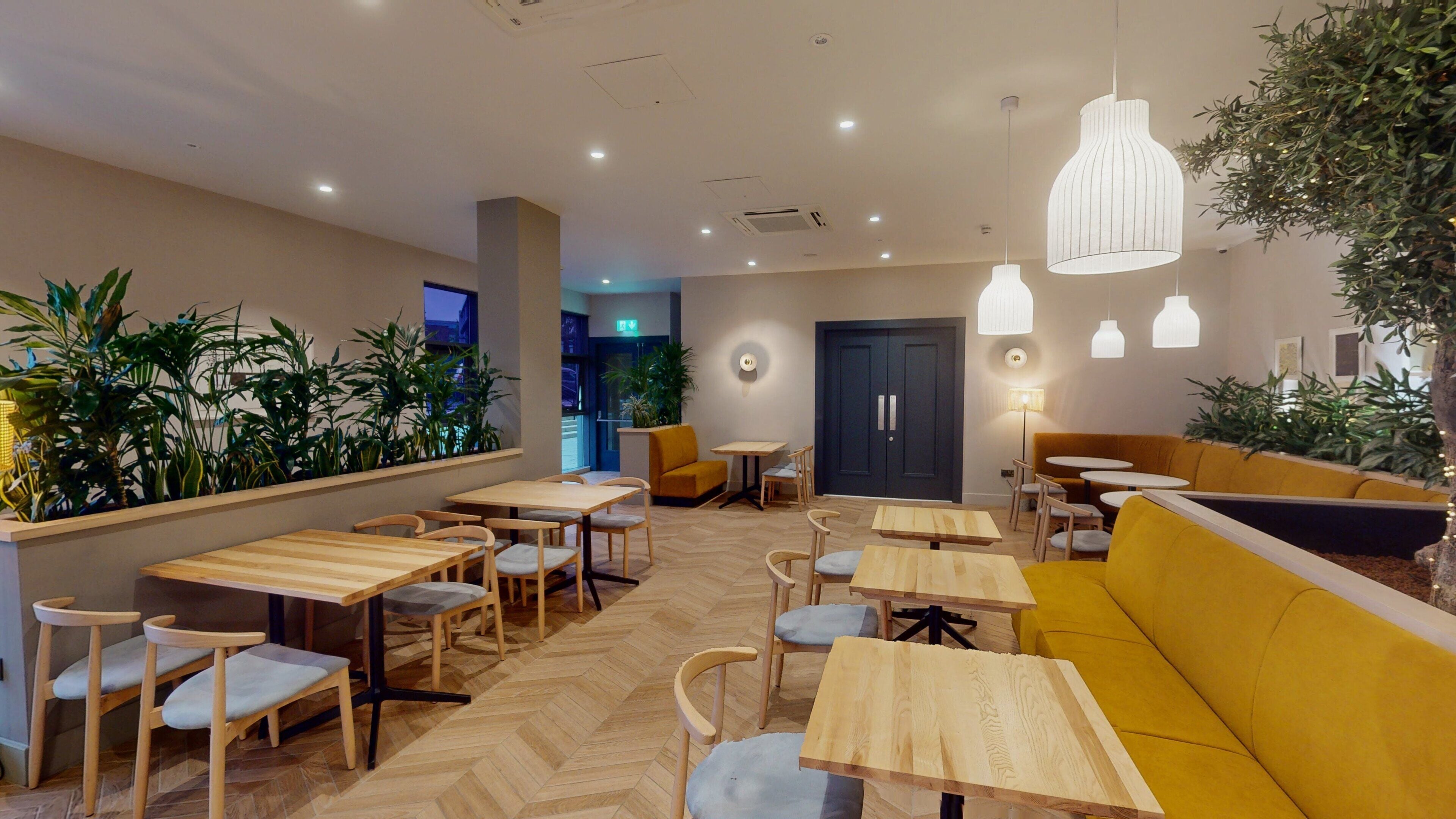 A modern indoor seating area with wooden tables and chairs, a yellow sofa, and potted plants.